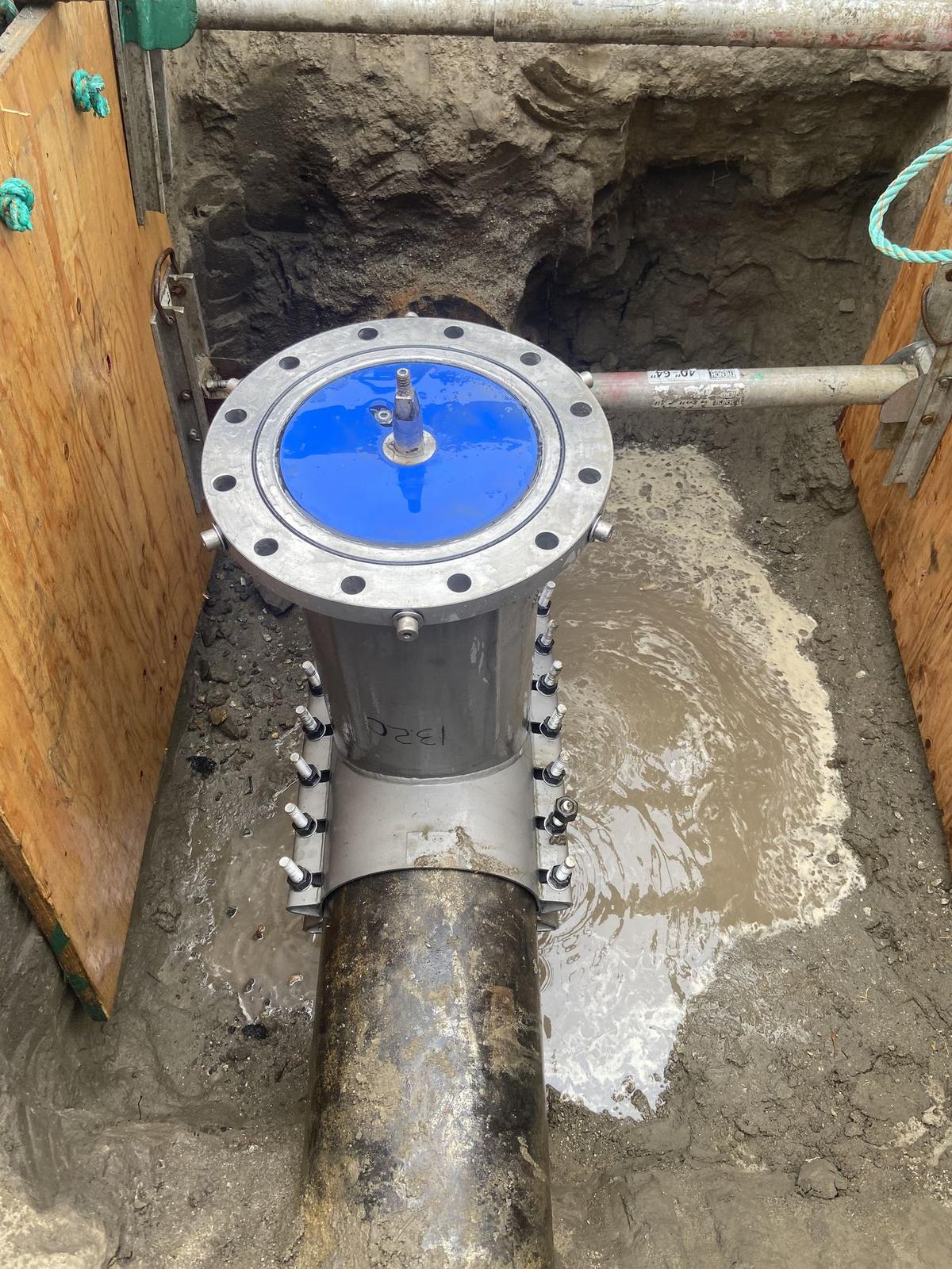Insertion valve body clamped to a water main in a muddy trench, blue cover bolted on top.