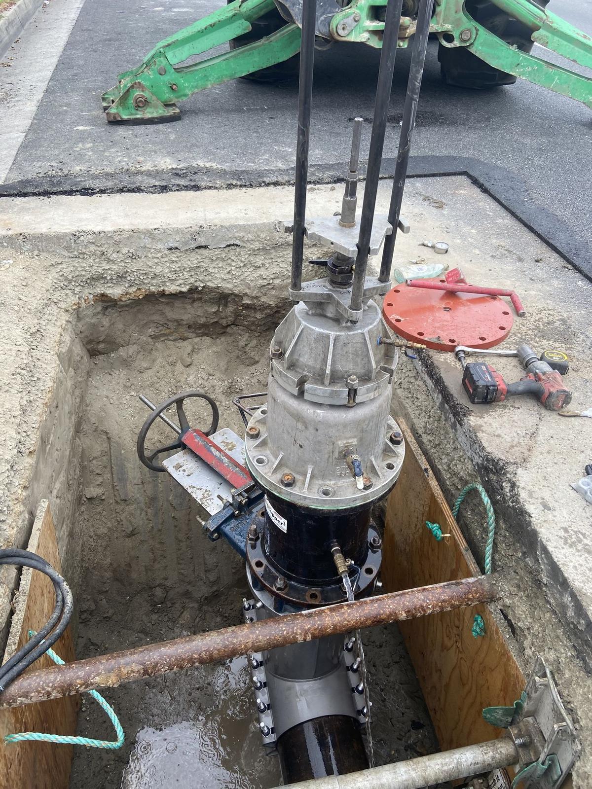 Tapping machine mounted on an insertion valve in a shored street excavation, with a backhoe at the curb.