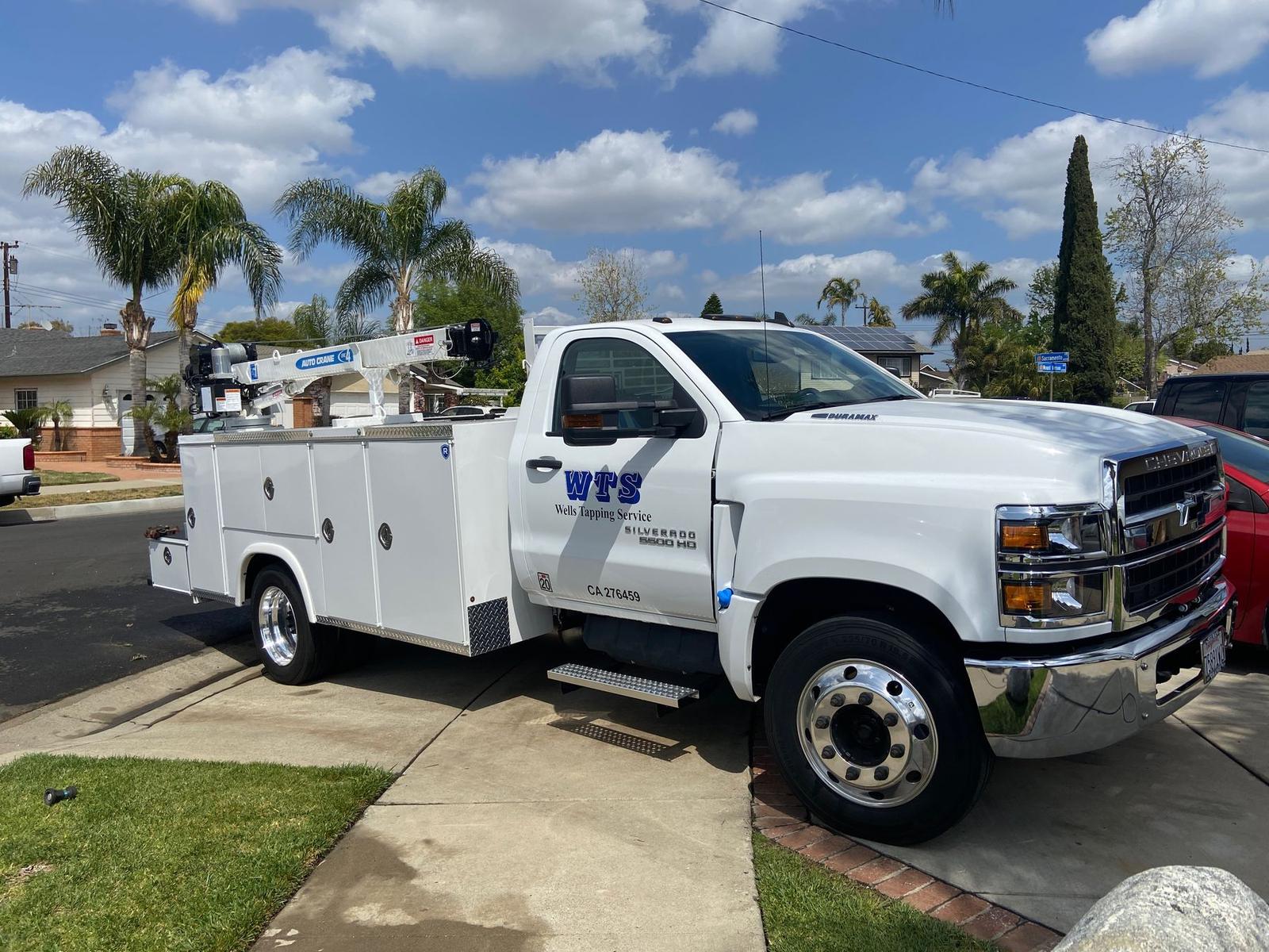White Chevrolet service truck with a crane, lettered WTS Wells Tapping Service, parked at the curb.